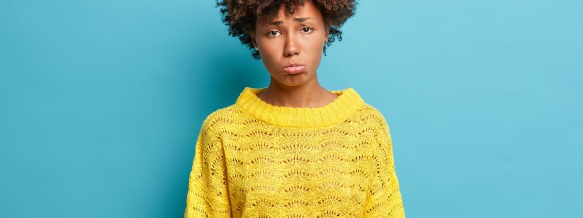 Attractive offended woman looks with sad frustrated expression purses lower lip and grimaces sadly at camera wears yellow knitted sweater isolated on blue background. Upset dark skinned girl